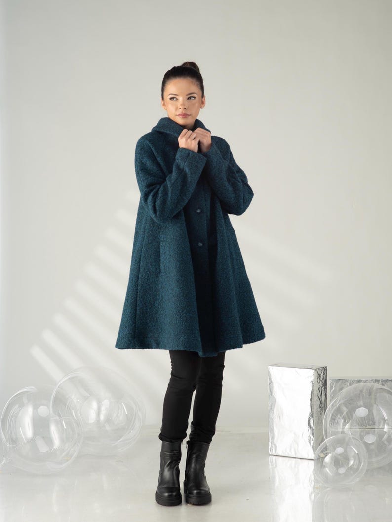 May include: A teal-colored, knee-length coat with a flared silhouette. The coat features a wide collar and button closure. The person in the image is wearing black pants and black boots. The coat is made of a textured fabric.