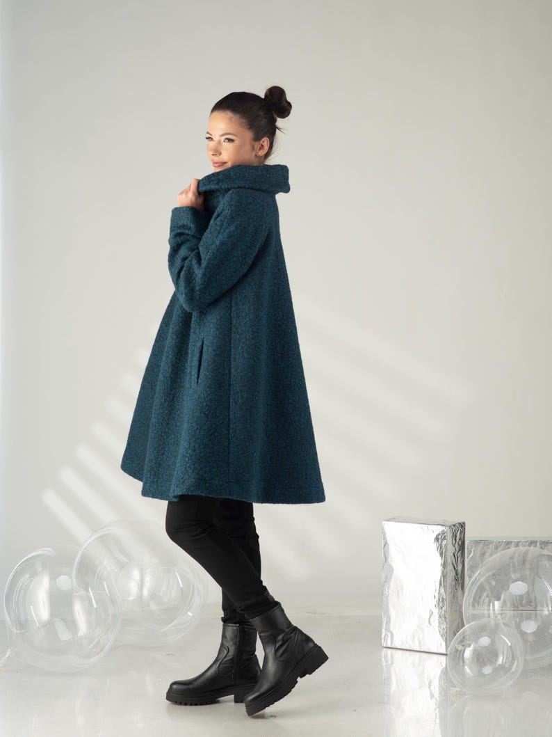 May include: A teal-colored, flared coat with a high collar. The coat is knee-length and has a textured appearance. The person is wearing black pants and black ankle boots. The background is a plain white.