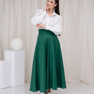 May include: A woman wearing a white button-down shirt with a green maxi skirt and white shoes. She is standing in front of a white backdrop.