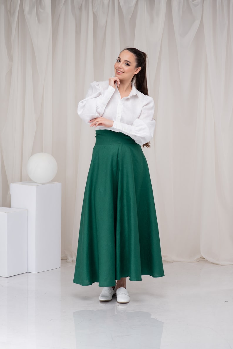 May include: A woman wearing a white button-down shirt with a green maxi skirt and white shoes. She is standing in front of a white backdrop.