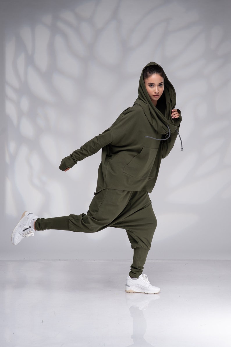 Puede incluir: Una mujer con una sudadera con capucha verde y pantalones a juego. La sudadera tiene un cuello de pico y los pantalones son holgados. Lleva zapatillas blancas.