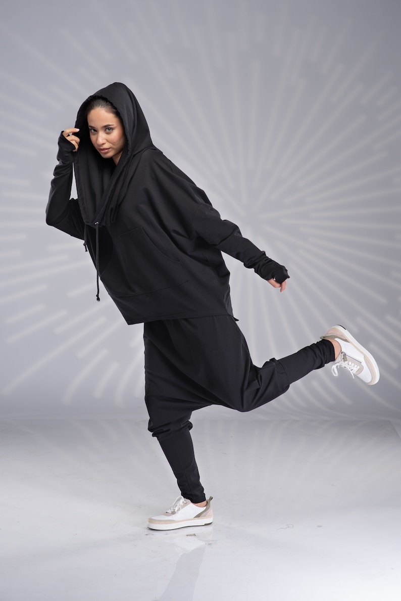 Puede incluir: Una persona con una sudadera con capucha negra y pantalones negros de corte holgado. La persona lleva zapatillas blancas y tiene la pierna derecha levantada.