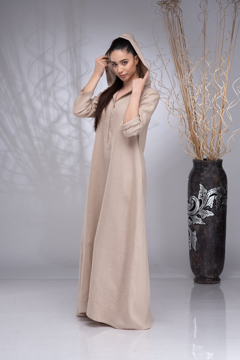 May include: A beige linen hooded dress with a zip fastening. The dress is long and flowing, and the model is wearing it with a casual, relaxed look.