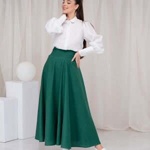 May include: A woman wearing a white collared blouse with puffed sleeves and a long, dark green skirt. She is standing in front of a white backdrop.