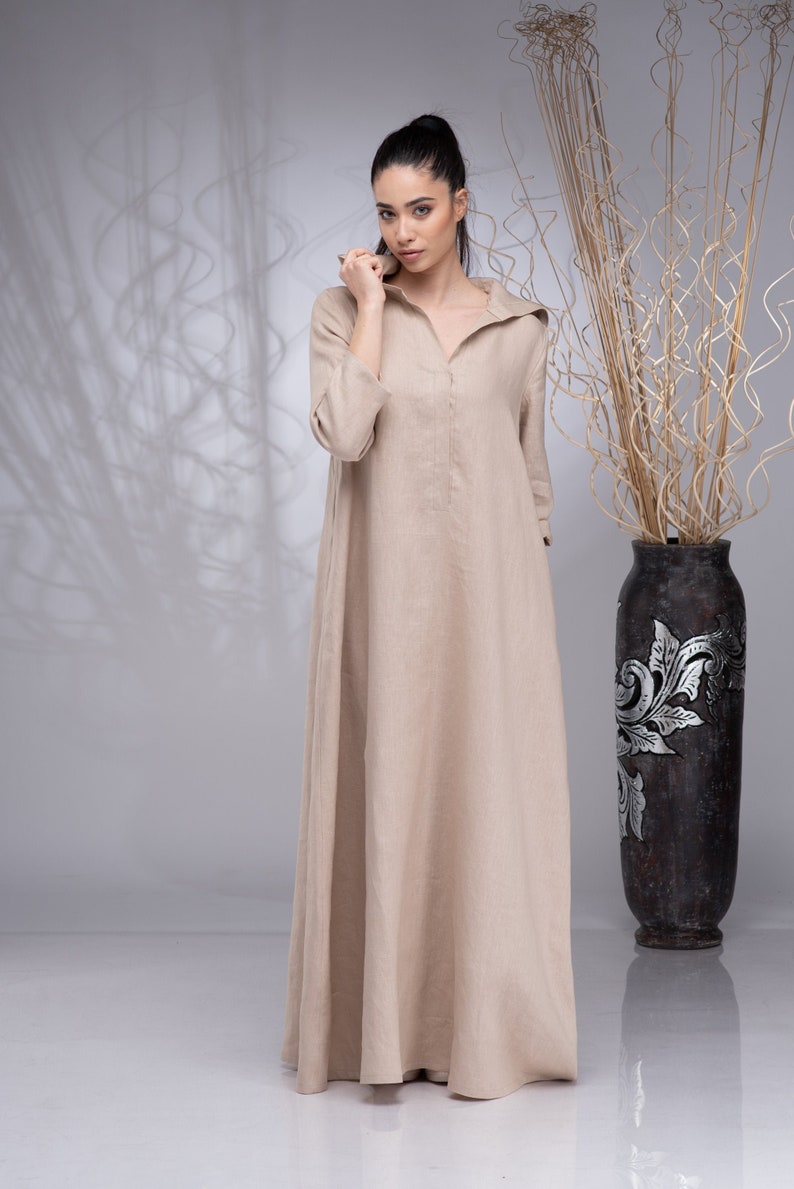 May include: A beige linen maxi dress with a collared neckline and long sleeves. The dress has a relaxed fit and a flowing silhouette.