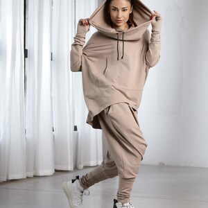 Puede incluir: Una mujer con una sudadera con capucha beige y pantalones a juego. La sudadera tiene cuello de pico y cord&oacute;n. Los pantalones son holgados y de pierna ancha.