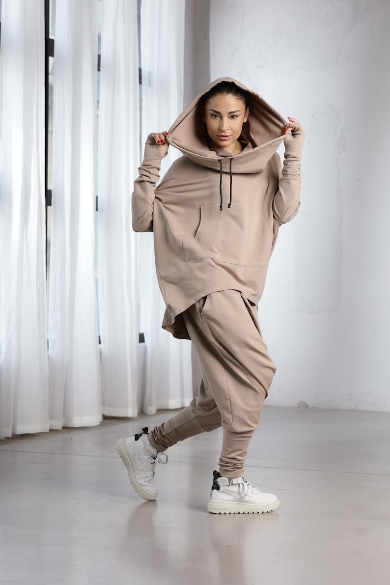 Puede incluir: Una mujer con una sudadera con capucha beige y pantalones a juego. La sudadera tiene cuello de pico y cord&oacute;n. Los pantalones son holgados y de pierna ancha.