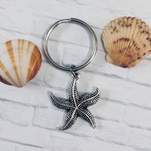 May include: A silver starfish keychain with a silver key ring and two seashells. The seashells are a light brown color with white stripes.