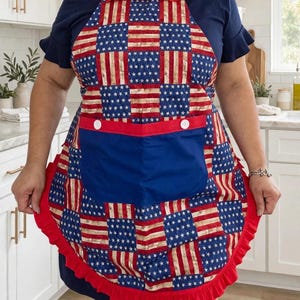 May include: An apron featuring a patchwork pattern of American flag motifs with alternating stars and stripes squares. The apron includes a solid blue front pocket with two white buttons, a red ruffled trim along the edges, and a red ruffled neckline.