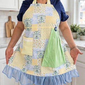 May include: A patchwork apron featuring a mix of yellow, blue, and white floral and checkered patterns. It includes a solid yellow pocket with white lace trim, a solid light green pleated panel attached with a blue button, and a light blue ruffled hem.