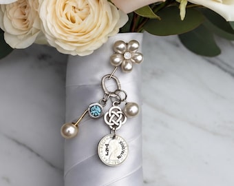May include: A bridal bouquet of cream, white, and dusty pink roses and ranunculus with green eucalyptus leaves. The stems are wrapped in silver satin ribbon, adorned with a silver charm featuring a small blue stone and a coin.