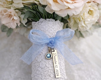 May include: A bridal bouquet featuring cream and blush roses. The stems are wrapped in white lace and tied with a light blue ribbon. A charm with a blue gemstone and the date "December 12, 2026" hangs from the ribbon.