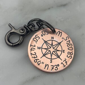 May include: A copper-colored round tag with a black compass design and coordinates. The tag is attached to a black metal clasp and ring. The compass rose is in the center, with the cardinal directions labeled. The coordinates are inscribed around the edge.