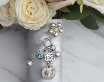 May include: A bridal bouquet of white and pale pink roses and ranunculus flowers with green eucalyptus leaves. The stems are wrapped in white satin ribbon, adorned with a silver charm featuring a flower, a blue stone, and a small coin.