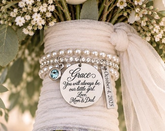 May include: A bridal bouquet handle wrapped in white fabric featuring a beaded bracelet with a round silver charm engraved with "Grace, You will always be our little girl. Love, Mom & Dad," a rectangular charm with "October 4, 2025," and a blue crystal.