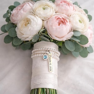 Pode incluir: Um buqu&ecirc; de noiva com flores rosa e brancas e folhagem verde. As hastes s&atilde;o envoltas em cetim e renda brancos, adornadas com pulseiras de p&eacute;rolas e um pingente com uma gema azul e a data "12 de dezembro de 2026".