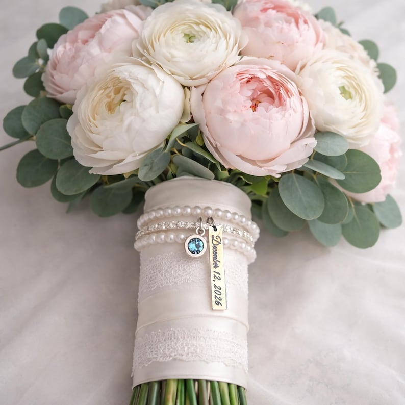 Pode incluir: Um buqu&ecirc; de noiva com flores rosa e brancas e folhagem verde. As hastes s&atilde;o envoltas em cetim e renda brancos, adornadas com pulseiras de p&eacute;rolas e um pingente com uma gema azul e a data "12 de dezembro de 2026".