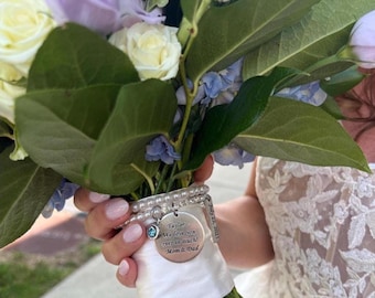 May include: A bridal bouquet featuring white and lavender roses, blue flowers, and lush green foliage. A silver charm bracelet with a round pendant and the inscription "Taylor We love you ever so much! Mom & Dad" is wrapped around the stems.
