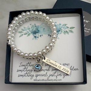 Pode incluir: Uma pulseira de p&eacute;rolas brancas com um berloque prateado e uma pedra preciosa azul, apresentada numa caixa de presente azul marinho. O berloque est&aacute; gravado com "December 12, 2026". O cart&atilde;o diz "Something new, something blue..." e o nome da loja Etsy.