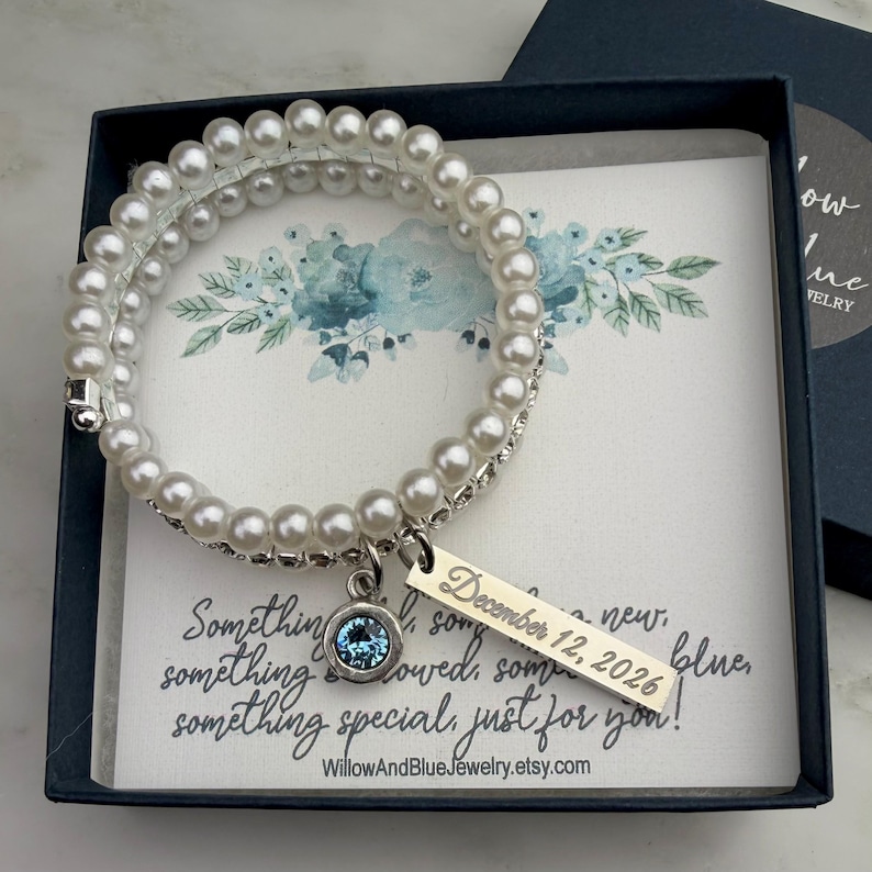 Pode incluir: Uma pulseira de p&eacute;rolas brancas com um berloque prateado e uma pedra preciosa azul, apresentada numa caixa de presente azul marinho. O berloque est&aacute; gravado com "December 12, 2026". O cart&atilde;o diz "Something new, something blue..." e o nome da loja Etsy.