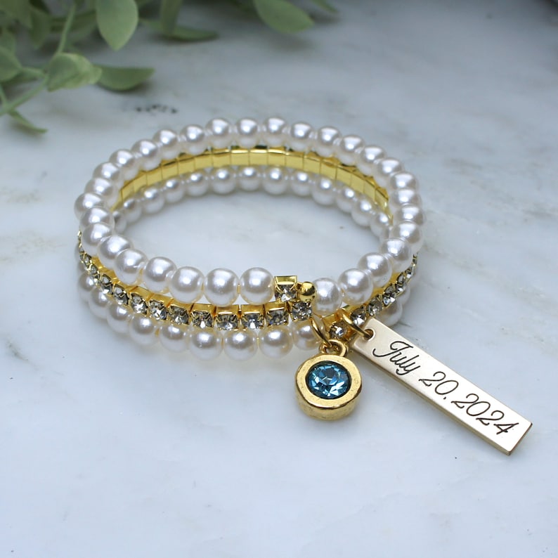 Pode incluir: Um conjunto de pulseiras empilh&aacute;veis com p&eacute;rolas falsas, detalhes em metal dourado e uma fileira de strass transparentes. Um berloque dourado com uma pedra preciosa azul e a inscri&ccedil;&atilde;o "July 20, 2024" pendura-se na pulseira.