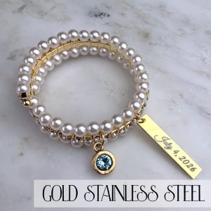 Pode incluir: Pulseira enrolada em a&ccedil;o inoxid&aacute;vel dourado com p&eacute;rolas falsas e um pequeno pingente de pedra preciosa azul. Uma etiqueta retangular dourada est&aacute; gravada com "July 4, 2026". A pulseira est&aacute; sobre uma superf&iacute;cie de m&aacute;rmore.