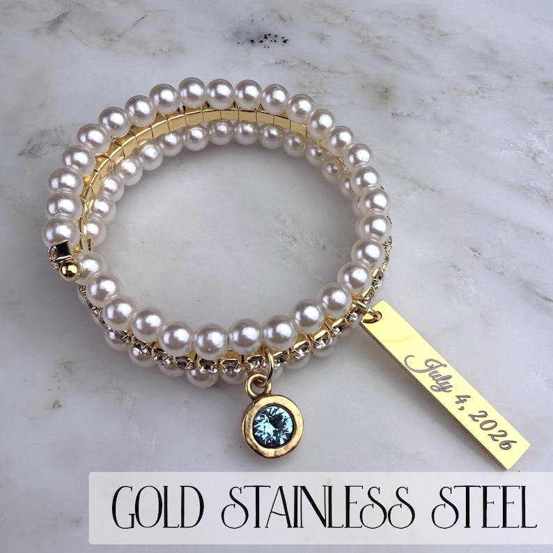 Pode incluir: Pulseira enrolada em a&ccedil;o inoxid&aacute;vel dourado com p&eacute;rolas falsas e um pequeno pingente de pedra preciosa azul. Uma etiqueta retangular dourada est&aacute; gravada com "July 4, 2026". A pulseira est&aacute; sobre uma superf&iacute;cie de m&aacute;rmore.