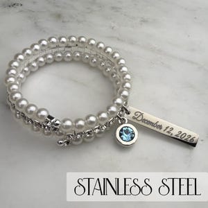 Pode incluir: Uma pulseira espiral com p&eacute;rolas de imita&ccedil;&atilde;o e uma fileira de strass transparentes. Um berloque prateado com uma pedra preciosa azul e uma etiqueta retangular gravada com "December 12, 2026" pendura-se na pulseira. O texto "STAINLESS STEEL" est&aacute; na parte inferior.