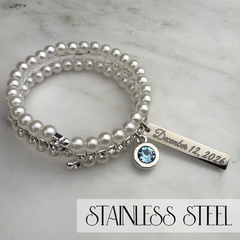 Pode incluir: Uma pulseira espiral com p&eacute;rolas de imita&ccedil;&atilde;o e uma fileira de strass transparentes. Um berloque prateado com uma pedra preciosa azul e uma etiqueta retangular gravada com "December 12, 2026" pendura-se na pulseira. O texto "STAINLESS STEEL" est&aacute; na parte inferior.