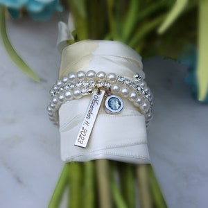 Pode incluir: Um buqu&ecirc; de noiva com uma al&ccedil;a envolta em fita creme. A al&ccedil;a &eacute; adornada com pulseiras de p&eacute;rolas e strass, e uma etiqueta de prata gravada com "November 11, 2023" e um berloque azul. O buqu&ecirc; inclui flores azuis e verdes.