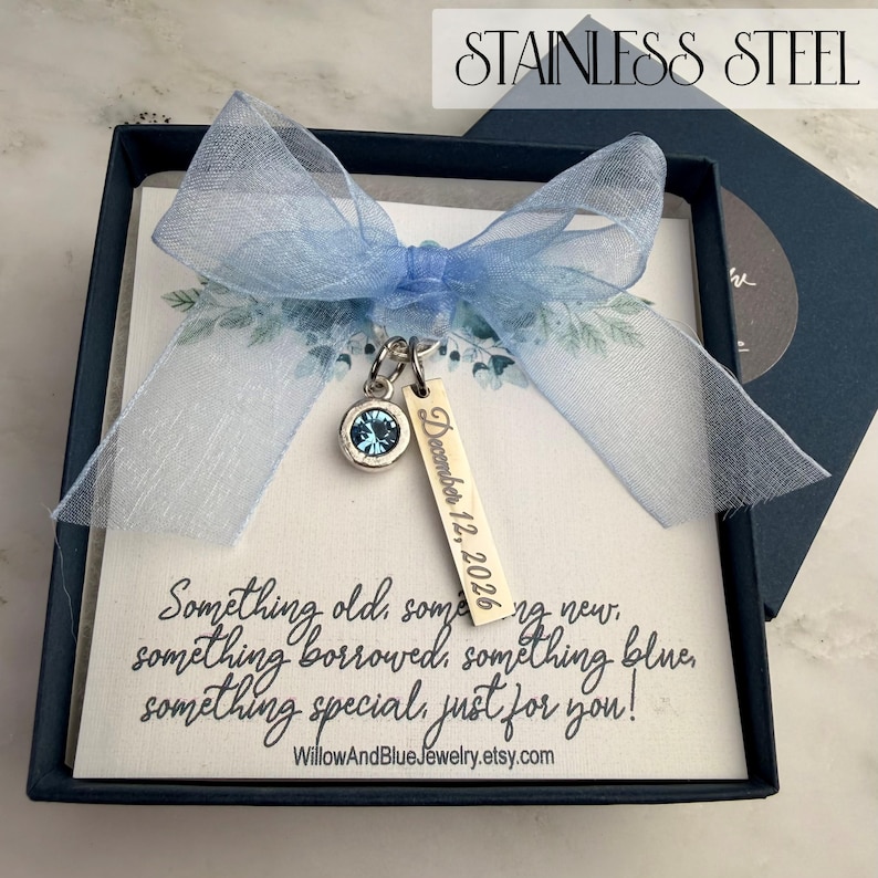 Op de afbeelding: Een marineblauwe geschenkdoos met een lichtblauw lint en een bedel. De bedel heeft een blauwe edelsteen en een label met de tekst "December 12, 2026". De kaart binnenin luidt "Something old, something new, something borrowed, something blue, something special, just for you!"