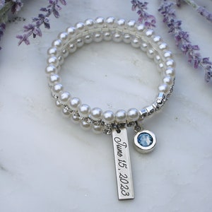 Pode incluir: Uma pulseira de p&eacute;rolas brancas com um berloque de prata que diz "June 15, 2023". A pulseira tamb&eacute;m tem um berloque de pedra preciosa azul. A pulseira est&aacute; em uma superf&iacute;cie branca com flores roxas.