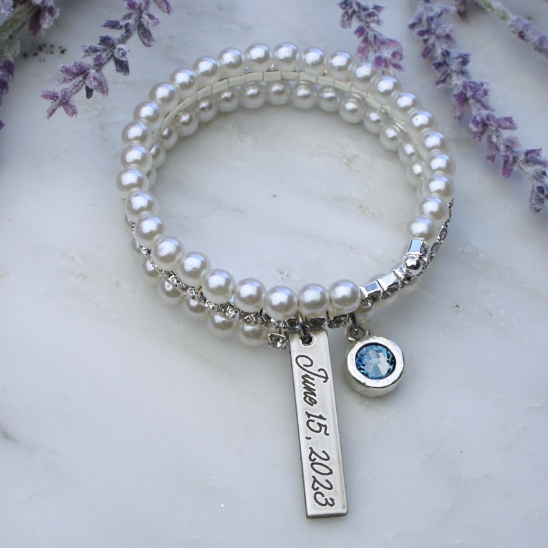 Pode incluir: Uma pulseira de p&eacute;rolas brancas com um berloque de prata que diz "June 15, 2023". A pulseira tamb&eacute;m tem um berloque de pedra preciosa azul. A pulseira est&aacute; em uma superf&iacute;cie branca com flores roxas.