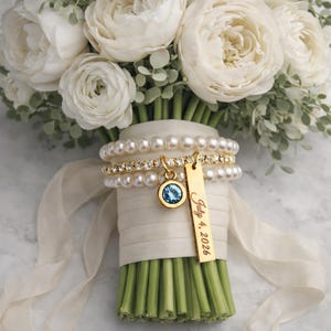 May include: A bridal bouquet of white flowers with green stems, wrapped with ivory ribbon. A gold charm with a blue stone and a gold tag with the inscription "July 4, 2026" are attached to the bouquet.