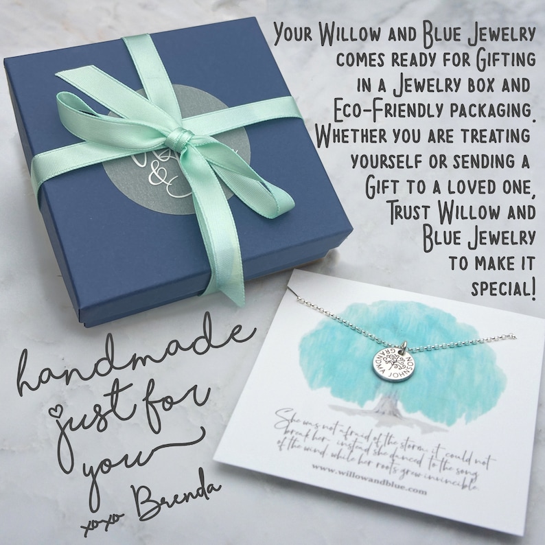 Op de afbeelding: Een marineblauwe sieradendoos met een lichtgroen lint, naast een zilveren ketting met een boomhanger. De doos heeft een cirkelvormig logo en de woorden "handmade just for you" zijn in cursief geschreven. De ketting ligt op een kaart met een boomillustratie en tekst.