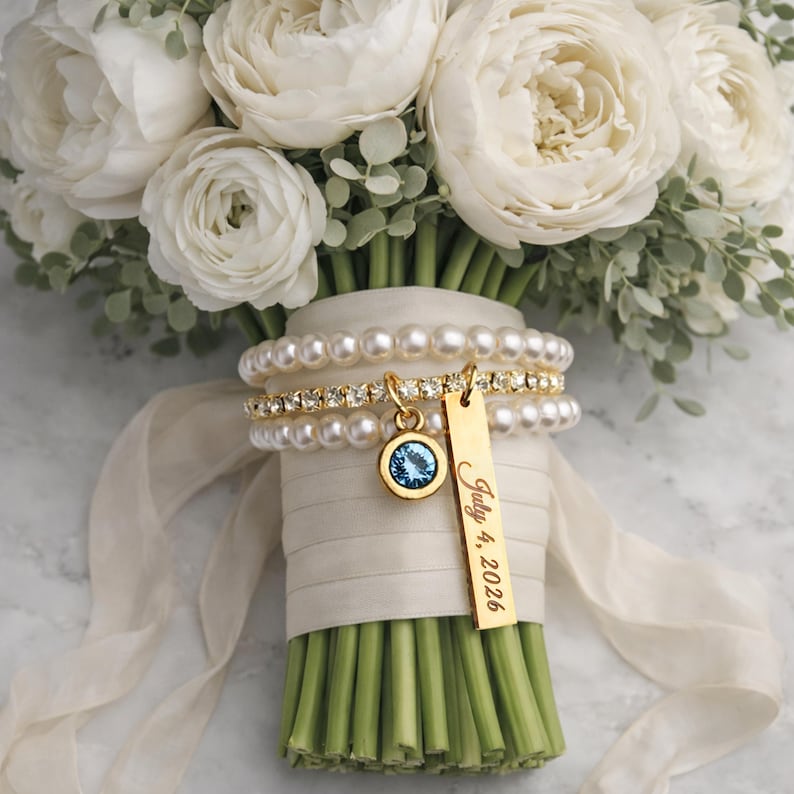 Pode incluir: Um buqu&ecirc; de noiva de flores brancas com caules verdes, envolto com fita marfim. Um pingente de ouro com uma pedra azul e uma etiqueta de ouro com a inscri&ccedil;&atilde;o "July 4, 2026" est&atilde;o anexados.