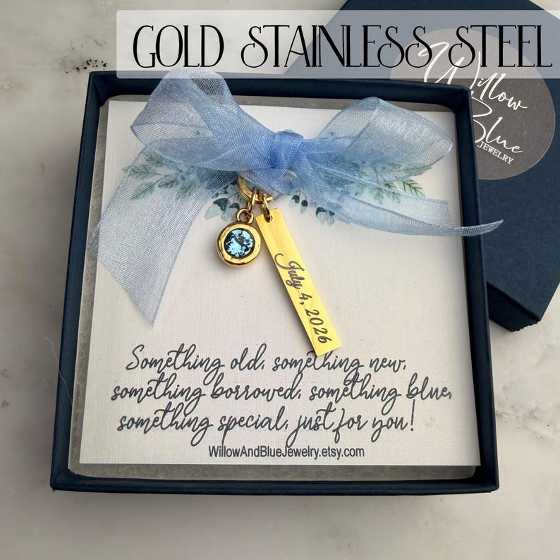 Op de afbeelding: Een gouden roestvrijstalen hanger met een blauwe edelsteen en een label met de inscriptie "July 4, 2026" hangt aan een blauw lint. De hanger wordt gepresenteerd in een marineblauwe doos met de tekst "Something old, something new..." en "WillowAndBlueJewelry.etsy.com".