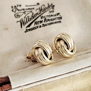 May include: A pair of polished gold stud earrings featuring a three-dimensional love knot design with smooth, rounded loops. The earrings are displayed inside a cream-coloured jewellery box with the text William Hardy 99 Victorian New Brighton.