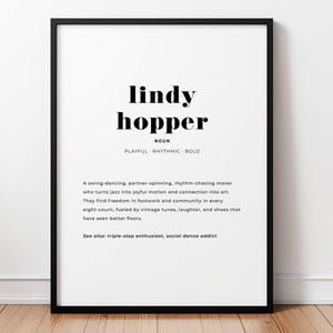 Puede incluir: Impresión enmarcada en negro con la palabra "lindy hopper" en negrita. La definición incluye "playful, rhythmic, bold" y una descripción de un bailarín de swing. La impresión está sobre un fondo blanco.