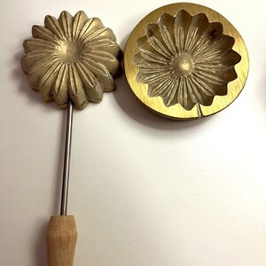 Op de afbeelding: Een vintage messing bloemvormige koekjesvorm en een bijpassend ijzer. De vorm is rond met een gedetailleerd bloemmotief. Het ijzer heeft een houten handvat en een vergelijkbare bloemvorm. De items worden waarschijnlijk gebruikt voor het maken van decoratieve koekjes.