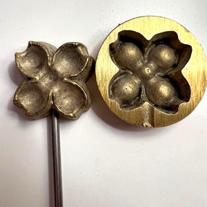 Könnte beinhalten: Zwei blumenförmige Küchenwerkzeuge aus Messing. Eines ist eine Form mit Griff, das andere eine kreisförmige Form. Beide Werkzeuge haben ein vierblättriges Blumendesign. Die Werkzeuge sind aus Messing und liegen auf einer weißen Oberfläche.