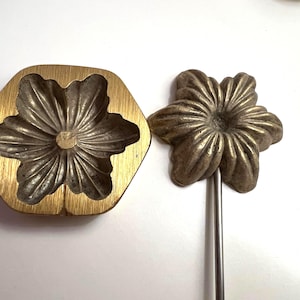 Op de afbeelding: Een goudkleurige, zeshoekige bloemvormige mal en een bronzen bloem op een metalen staaf. De bloem heeft gedetailleerde bloemblaadjes. Deze items zijn waarschijnlijk voor knutsel- of decoratieve doeleinden. De mal is ongeveer 10 cm breed.