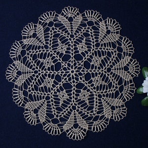 Può includere: Un delicato tovagliolo all'uncinetto bianco con un design circolare, che presenta intricati motivi floreali. Il tovagliolo è mostrato su uno sfondo blu scuro.