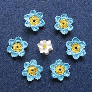 May include: A collection of handmade crochet flowers in a circular arrangement. The flowers have light blue petals with yellow centers. A single white flower with a yellow center is also present. The background is a solid blue fabric.