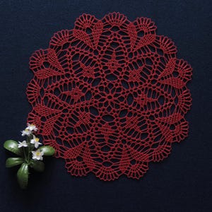 May include: A handmade, red crocheted doily with a detailed, openwork design. The circular doily is placed on a dark blue surface, with a small sprig of white flowers in the lower left corner. This decorative piece adds a touch of vintage charm.
