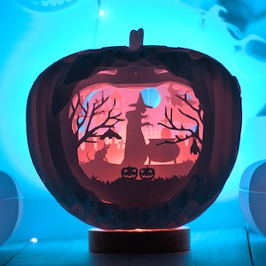 May include: A papercraft pumpkin with a glowing scene inside. The scene features a witch, a cauldron, a cat, and a full moon. The pumpkin is lit from within, casting a warm glow.