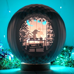 May include: A white paper sphere with a cut-out design of Santa's workshop. The sphere is lit from inside, casting a warm glow. The sphere is sitting on a wooden base and surrounded by green tinsel and disco balls.