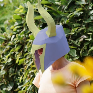 Puede incluir: Un casco de papel azul y amarillo con cuernos curvos. El casco lo lleva una persona, con una camisa amarilla visible. El fondo presenta follaje verde y flores amarillas.