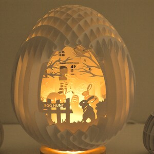 May include: A white paper egg-shaped lamp with a cut-out design of a house, trees, and bunnies. The lamp is lit from inside, casting a warm glow. The text "Egg Hunt" is visible on the front of the lamp.