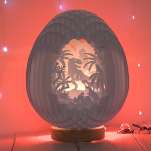 Può includere: Una lampada a forma di uovo bianco in cartapesta con un design a taglio di un dinosauro in una scena di giungla. La lampada è illuminata dall'interno, proiettando un bagliore caldo.